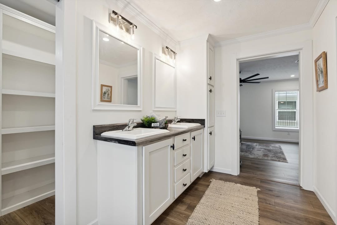 Modern bathroom with dual sinks, white cabinets, and two large mirrors. Soft lighting creates a clean, airy feel. Open door reveals a bedroom.