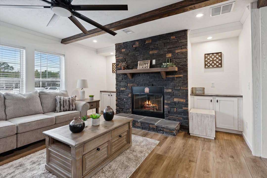 Cozy living room with a stone fireplace, lit fire, and a "Happy Place" sign. Gray sofa, wooden table, and decorative plants create a warm, inviting atmosphere.