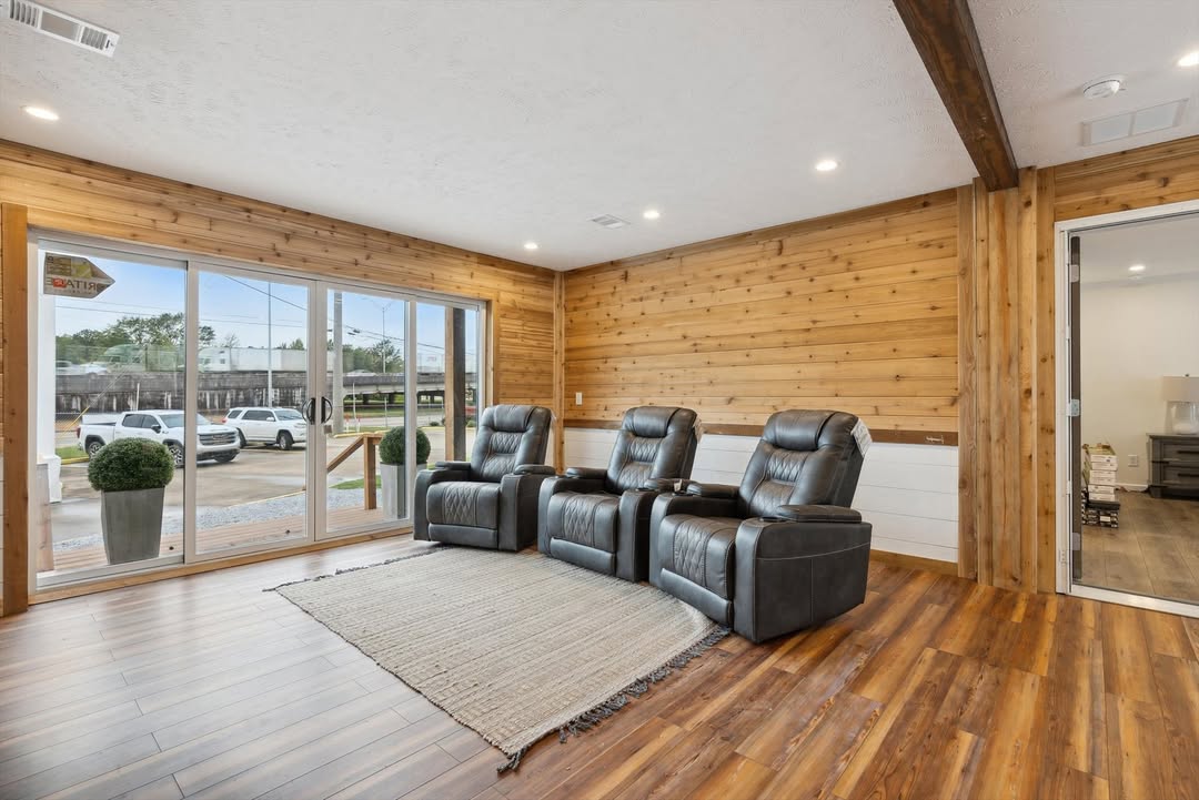 Modern living room with four black recliners on a light rug, wooden walls, and floor-to-ceiling windows. A calm, cozy atmosphere is conveyed.