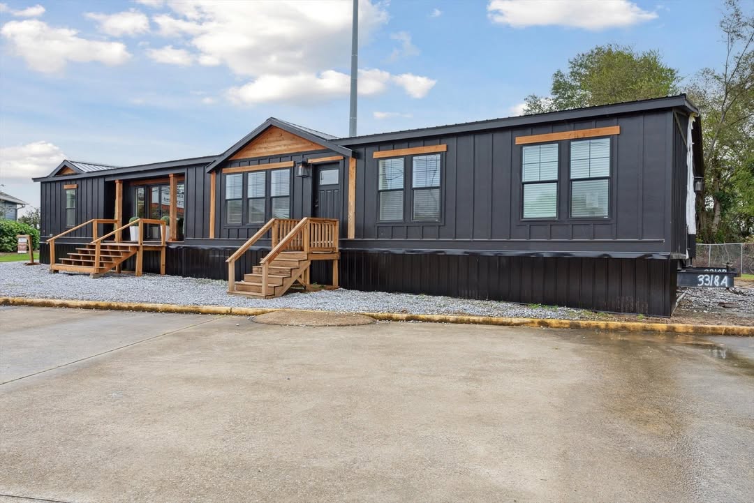 A modern, dark gray modular home with a wooden porch and stairs sits on a gravel lot. There's a clear blue sky with scattered clouds overhead.