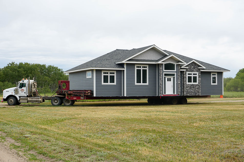 A large truck hauls a gray, fully constructed house on a wide trailer across a grassy field. The sky is overcast, with lush green trees in the background.