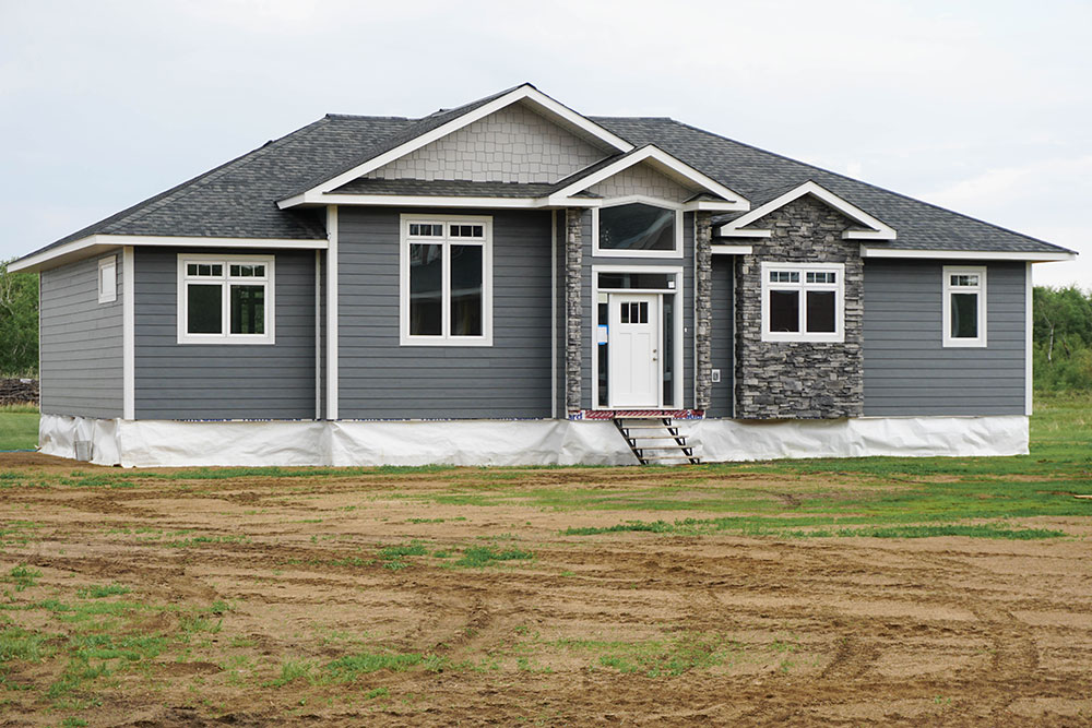 Single-story, gray modular home with white trim and stone accents. Set on grassy, undeveloped land under an overcast sky, creating a serene tone.