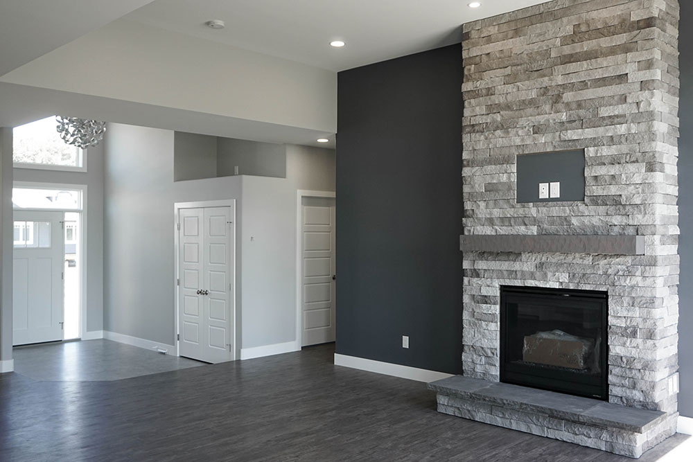 A modern living room with gray wooden floors and high ceilings. A large stone fireplace on the right contrasts with light gray walls, creating a sleek and inviting ambiance.