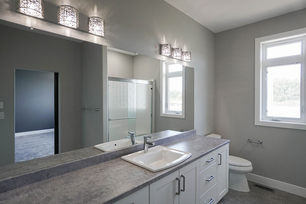 Modern bathroom with gray walls, double sinks, and a large mirror. Decorative lights above, window to the right, shower and toilet visible. Calm ambiance.