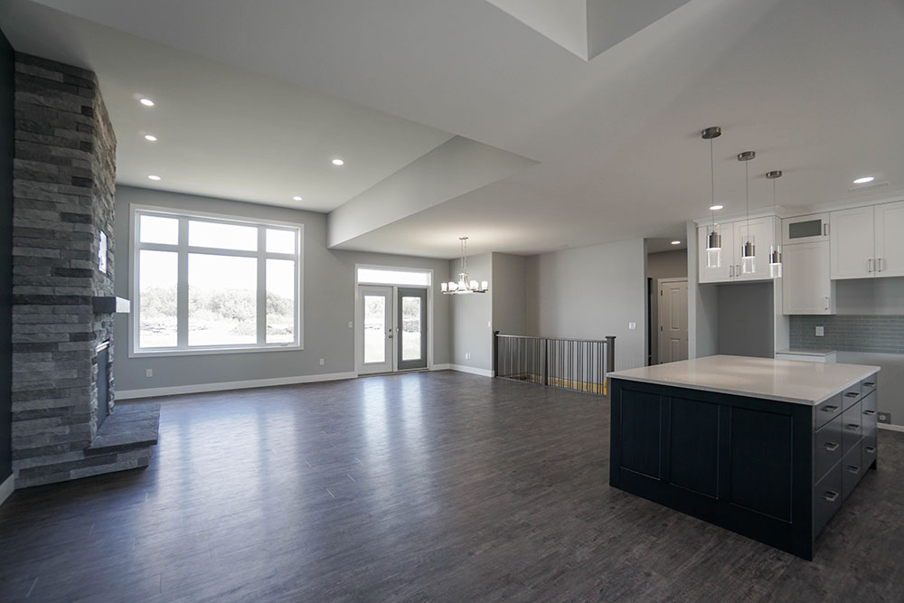 Spacious modern living area with dark wood flooring, large windows, a stone fireplace, white kitchen cabinets, and a central island under pendant lights.