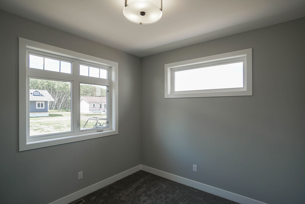 A small, empty room with gray walls features two windows; one large with a view of houses and trees, and a smaller one above. Soft light from a ceiling fixture.