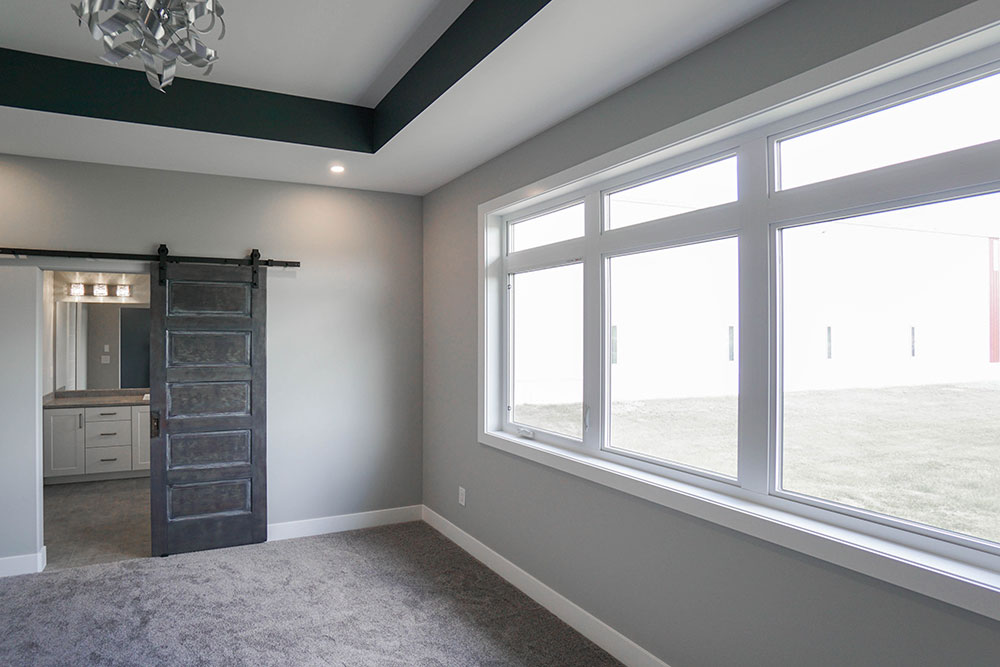 Modern room with grey walls, featuring a large window on the right, soft carpet, and a dark sliding barn door leading to a bright bathroom.