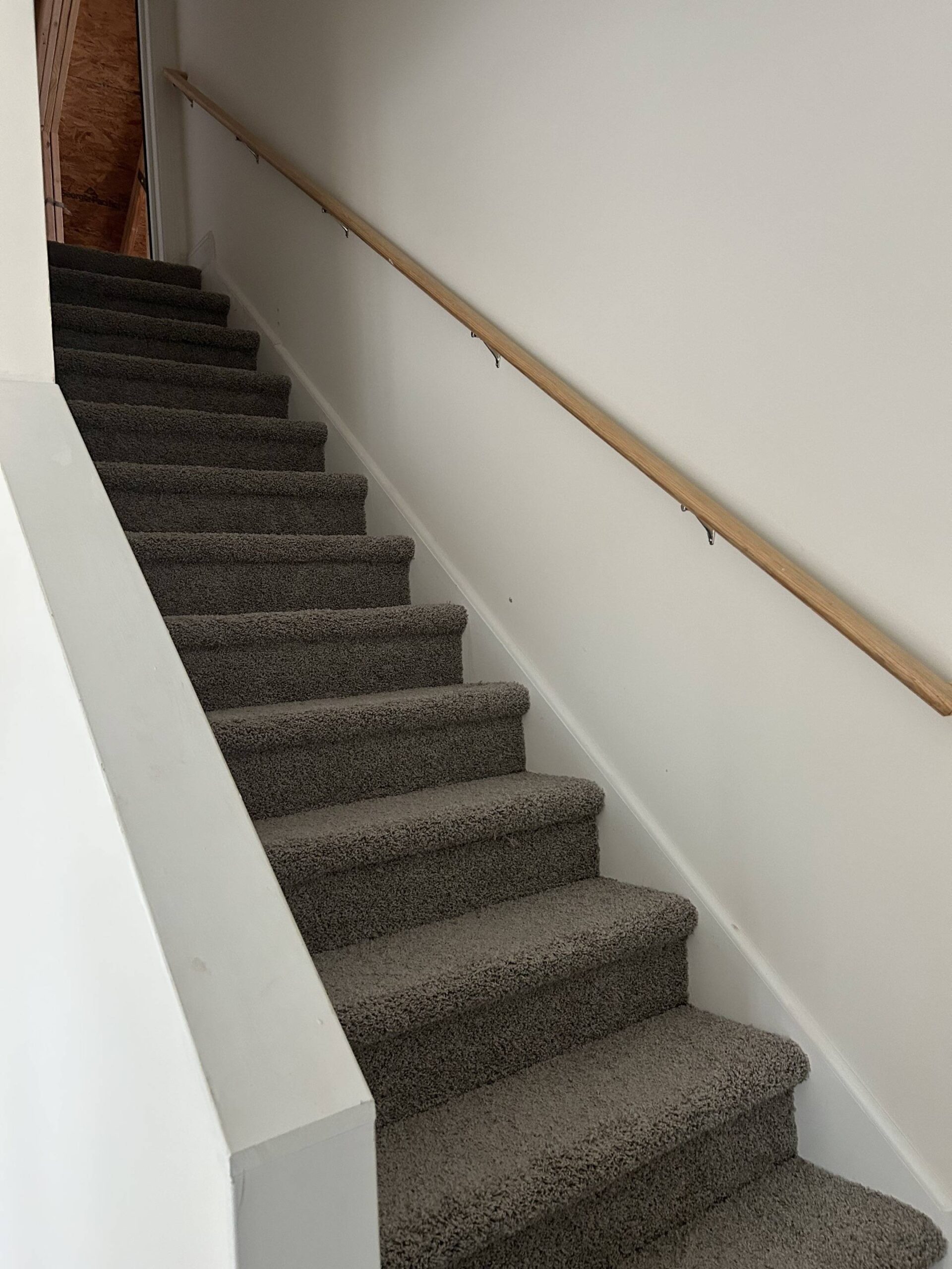 A carpeted staircase with a wooden handrail on the right ascends in a quiet, well-lit indoor space. The walls are white, creating a clean, minimalistic feel.