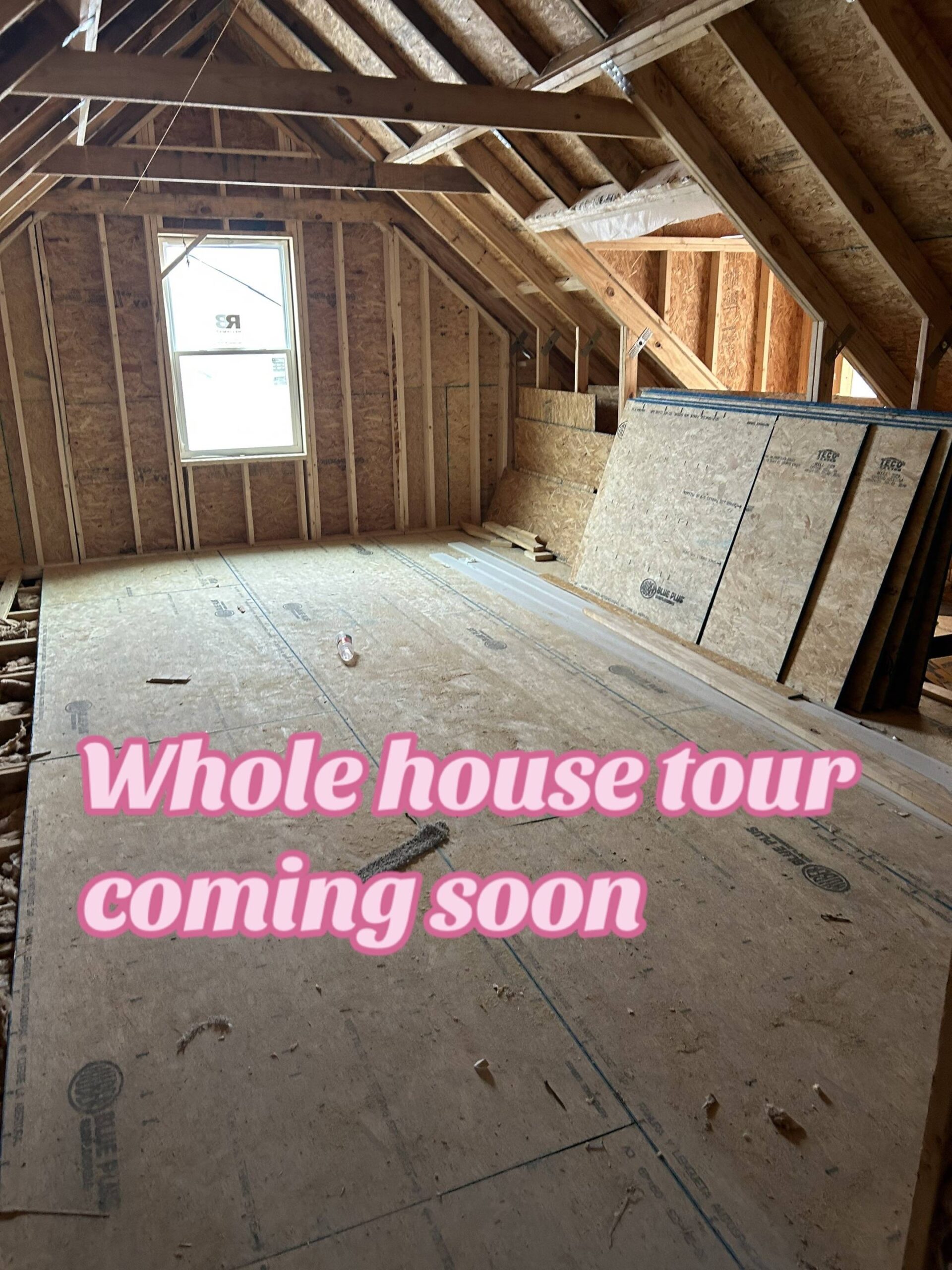Unfinished attic space with exposed wooden beams and a single window. Plywood covers the floor, with stacked boards on the right. Text reads "Whole house tour coming soon" in pink. Atmosphere is anticipatory.