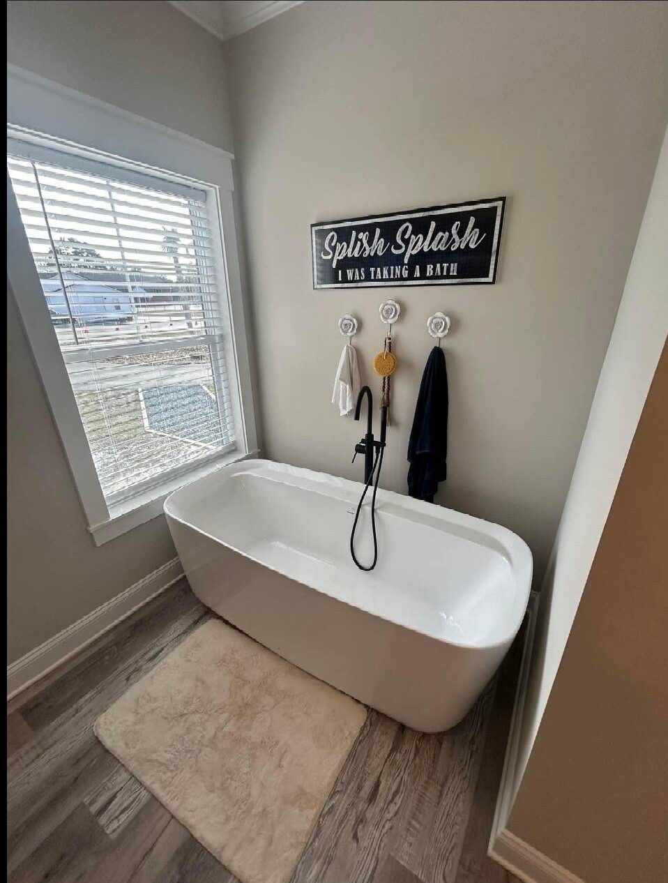 Modern bathroom with a white freestanding tub, beige rug, and wall hooks holding towels. A window with blinds and a sign saying “Splish Splash” add charm.