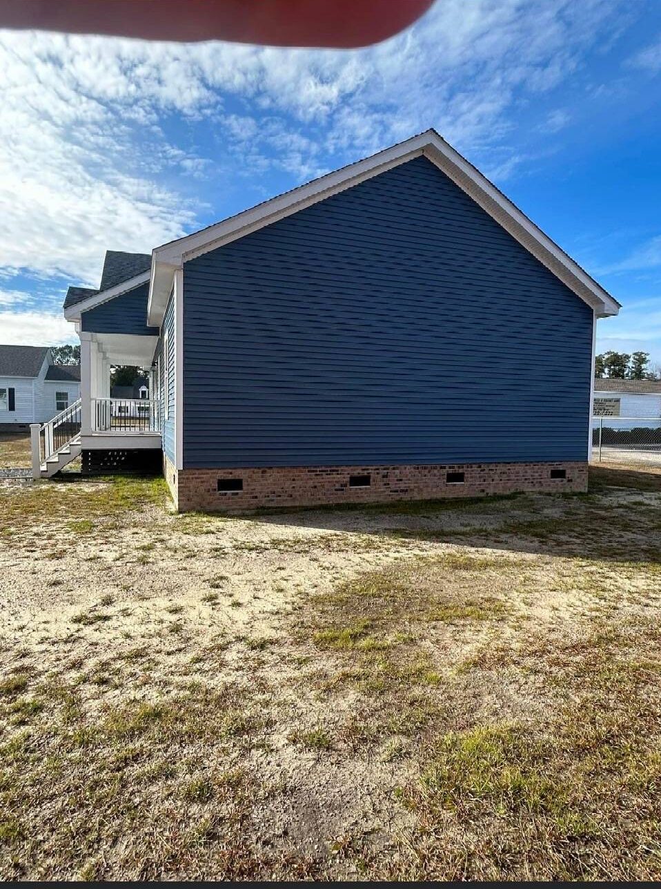 A blue house with a pitched roof, situated on a sandy, grassy lot under a partly cloudy sky. The side view shows white trim and a brick foundation.