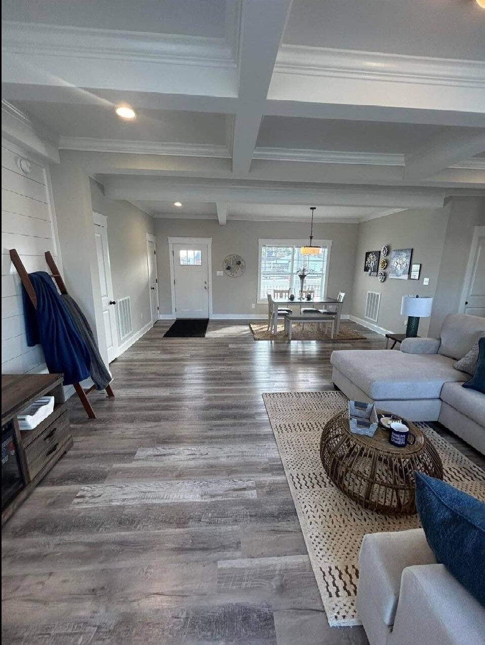 A bright, open living room features a large beige sectional sofa on a woven rug, a wooden coffee table, dining area in the background, and modern décor.