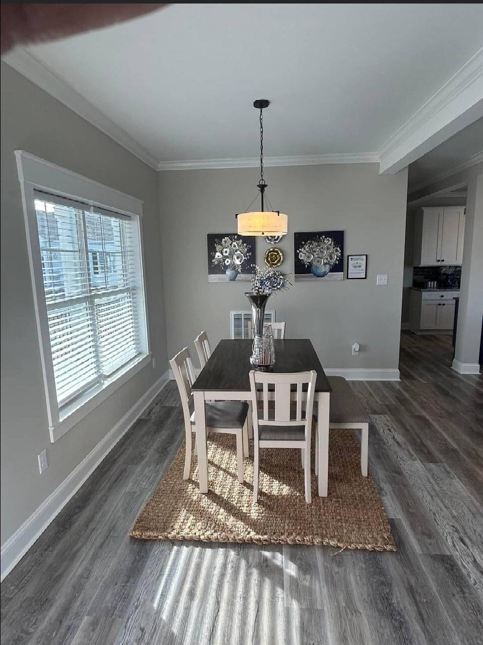 Bright dining room with a wooden table, white chairs, and a bench on a woven rug. A central pendant light hangs above, with artwork on the gray walls.