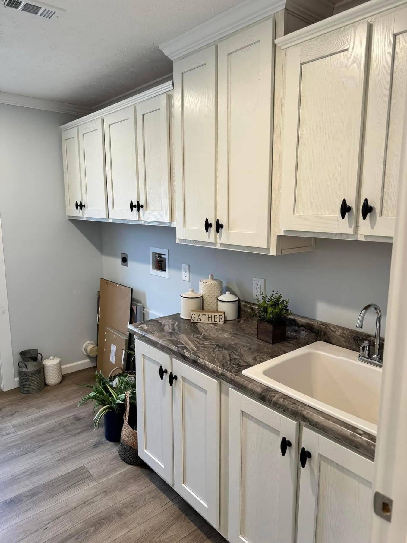 Laundry room with white cabinets and a marble countertop. A sink is on the right, and decor includes a "Gather" sign and small potted plants. Cozy and organized.