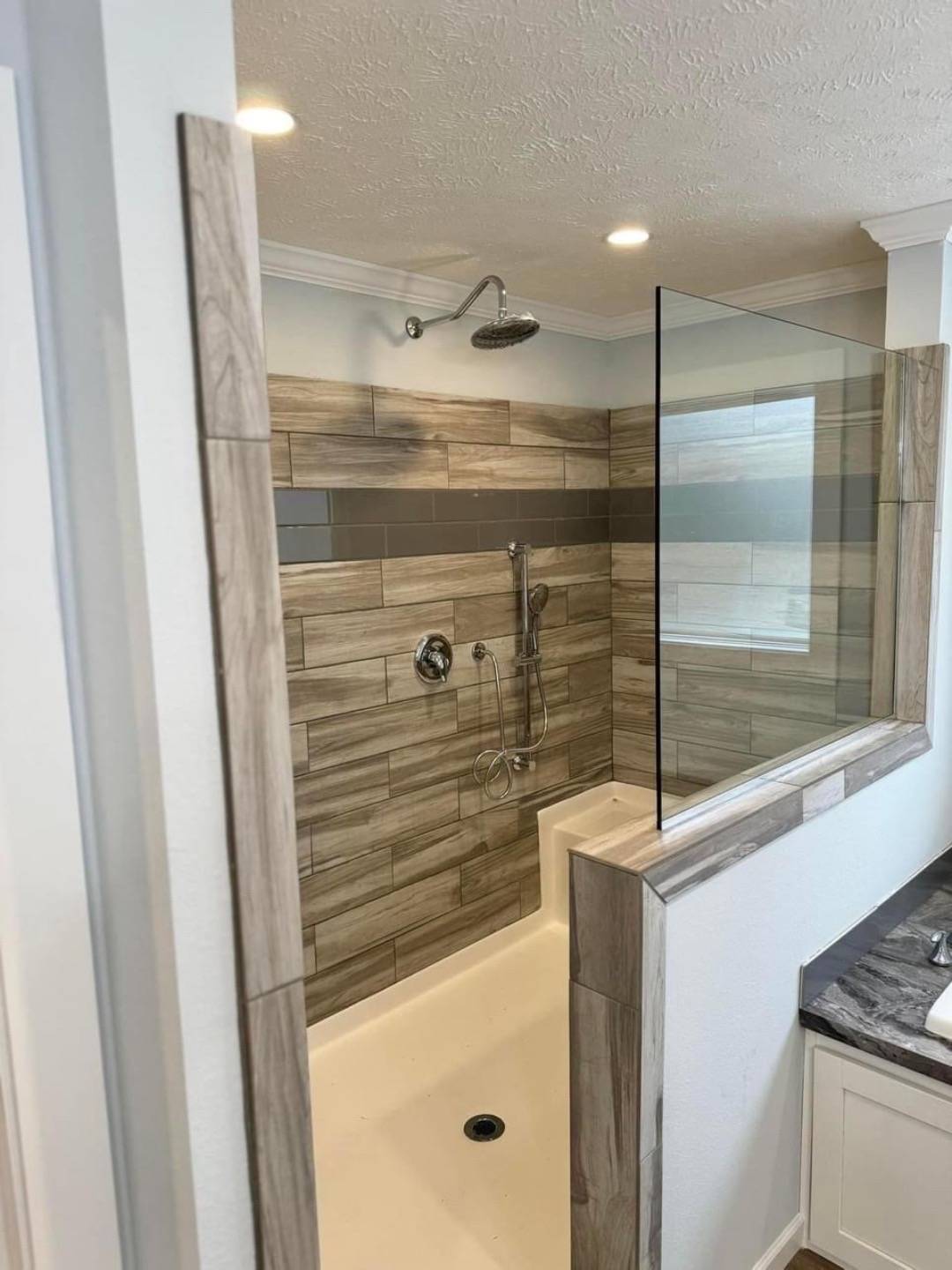 Modern bathroom with a shower and tub combo. Beige tiles with a dark accent stripe. Glass partition and overhead shower, creating a sleek, clean appearance.