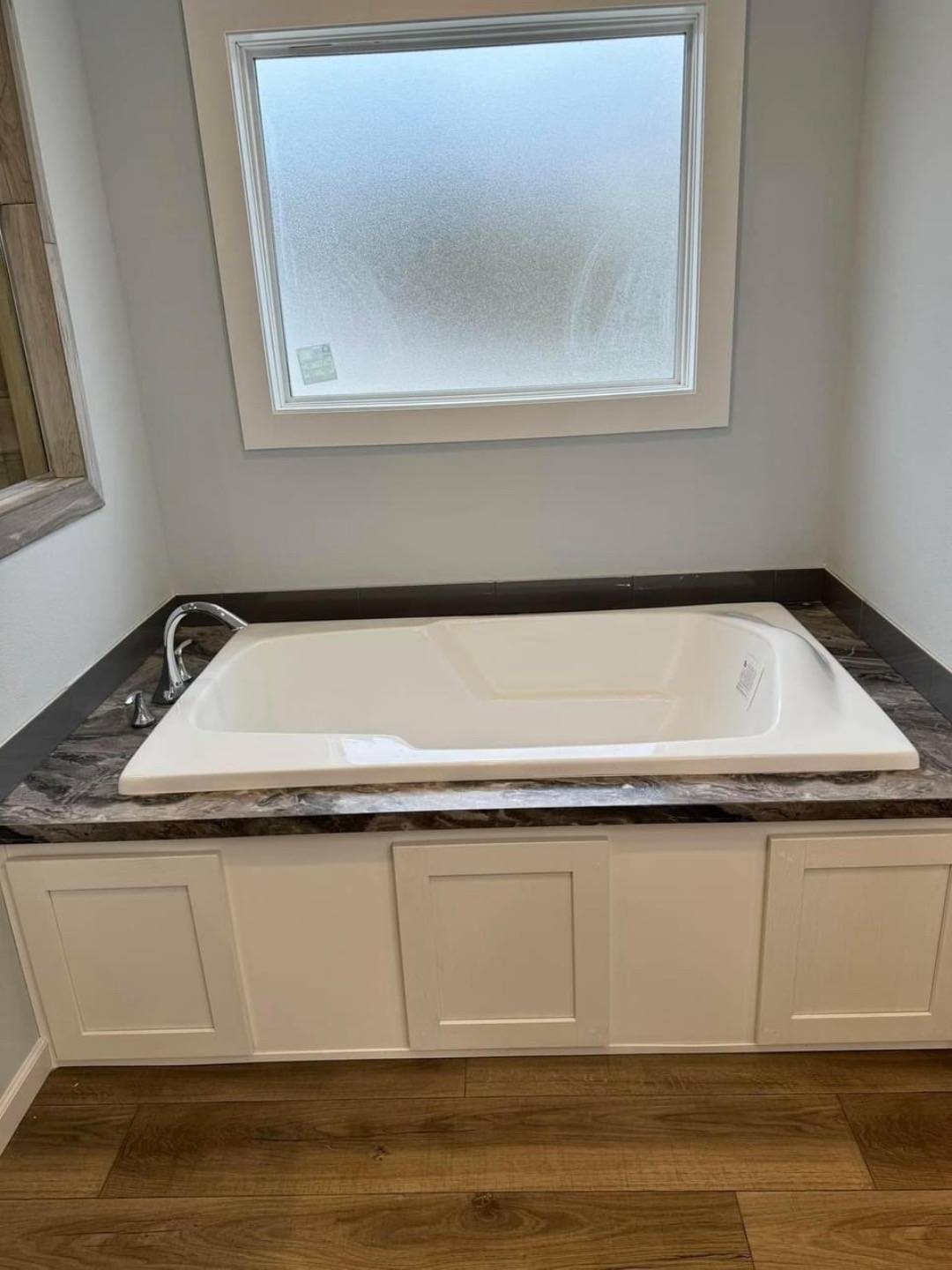 A pristine white bathtub is set in a marble countertop beneath a frosted window. The setting is a neat bathroom corner with a light, calming tone.
