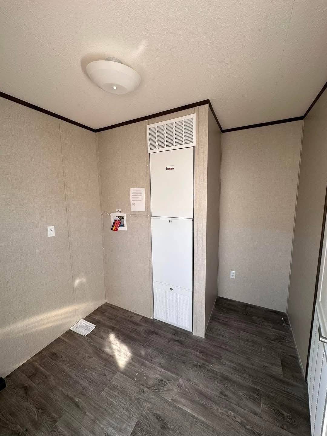 Compact room with beige walls and dark wood laminate flooring. Central heating unit is installed in the corner, with papers and a fire alarm nearby. Ceiling light fixture above.