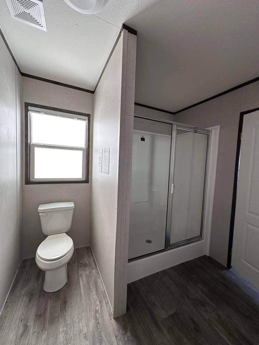 Minimalist bathroom with a white toilet by a small window and a sliding glass shower. The dark wood floor and muted walls create a clean, modern feel.