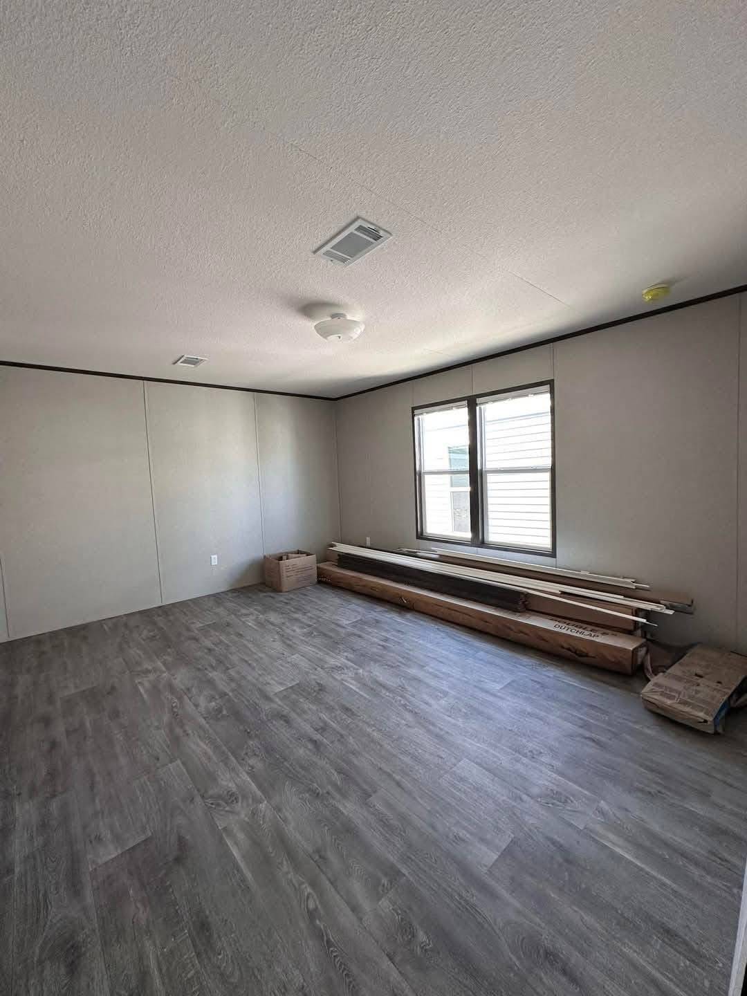 An empty room with gray wood flooring and beige walls. A window lets in natural light. Lumber and cardboard boxes are stacked against a wall.
