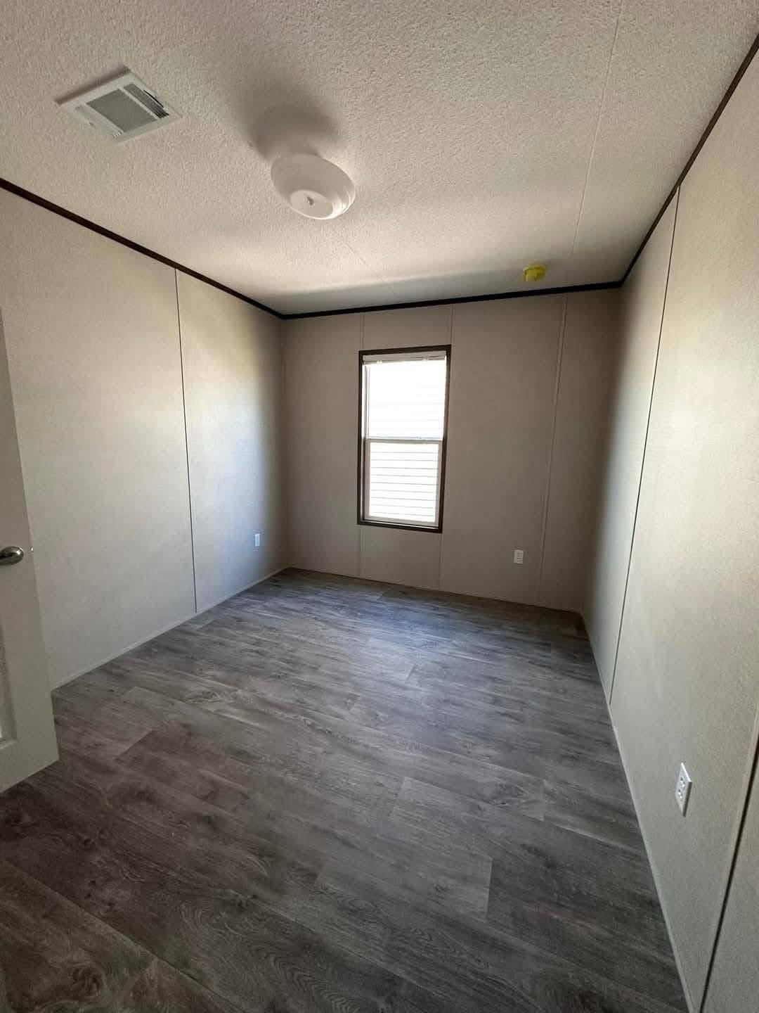 An empty room with beige walls and wood-textured laminate flooring. A single narrow window lets in light, and a ceiling light fixture is visible. Minimalist and clean.