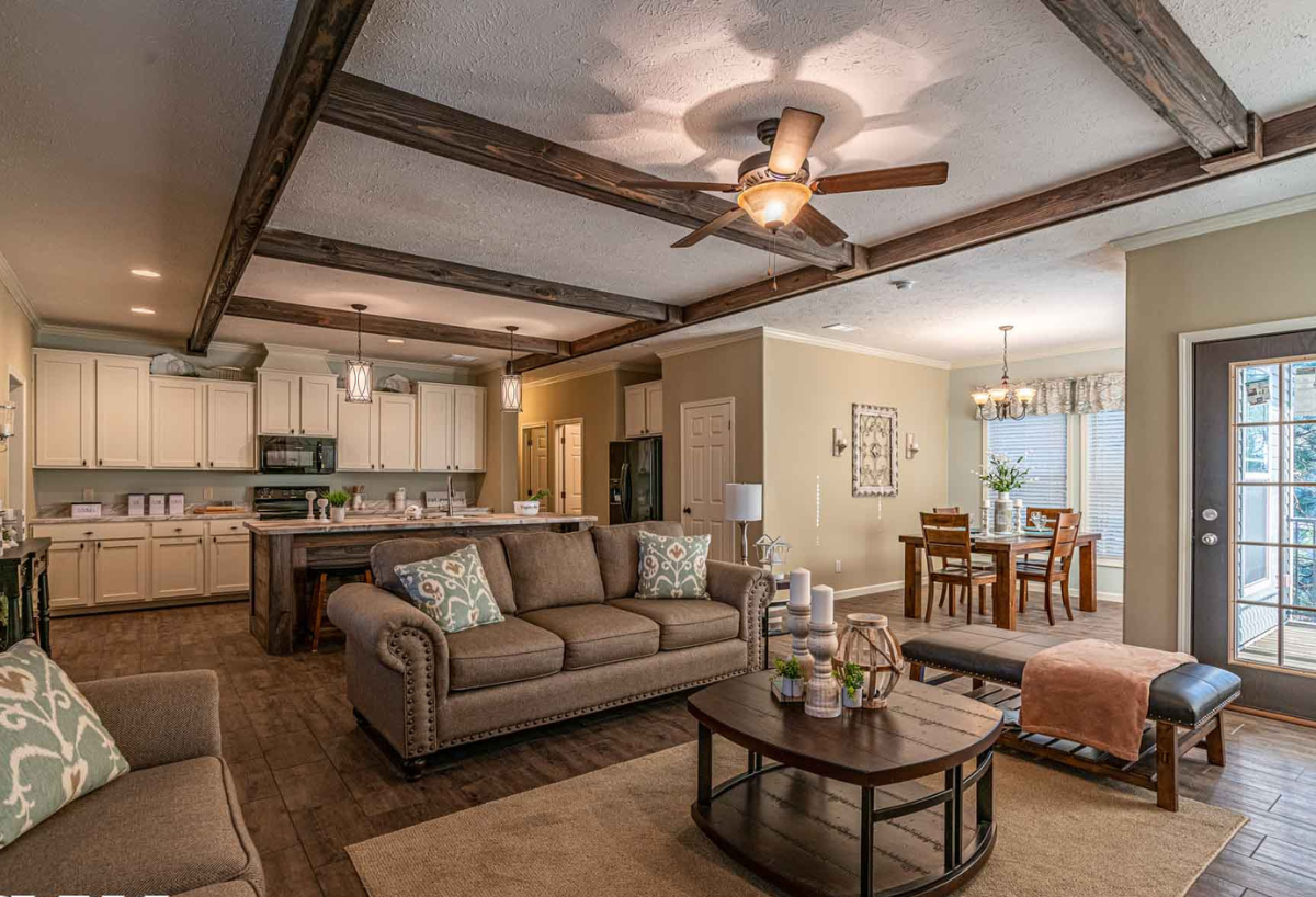 Cozy open-plan living room with beige sofas, a wooden coffee table, and ceiling fan. Adjacent is a kitchen with white cabinets and a dining area. Warm, inviting ambiance.