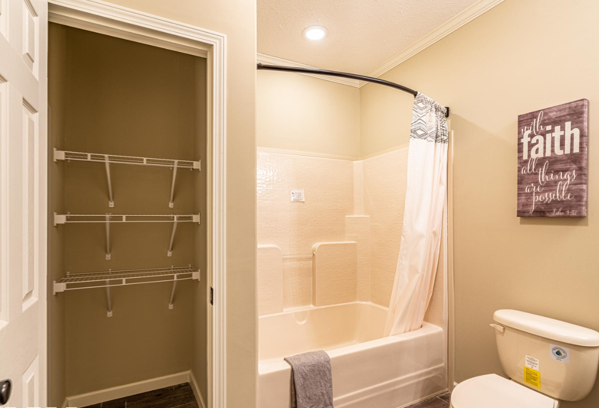 Bathroom with a beige bathtub and shower curtain, adjacent empty shelving, and a toilet. A sign saying "Faith" hangs on the wall, creating a serene atmosphere.