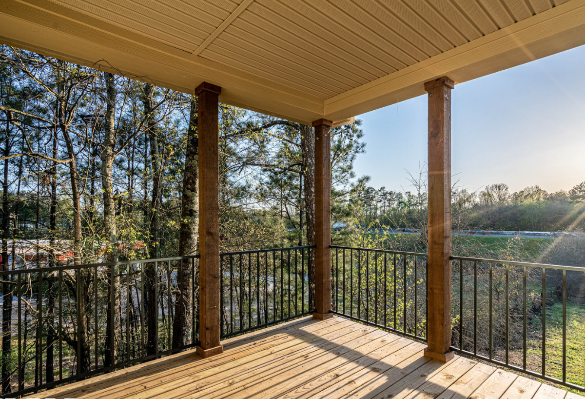 Covered wooden balcony with metal railings surrounded by trees, overlooking a peaceful, sunlit landscape. The setting conveys tranquility and warmth.