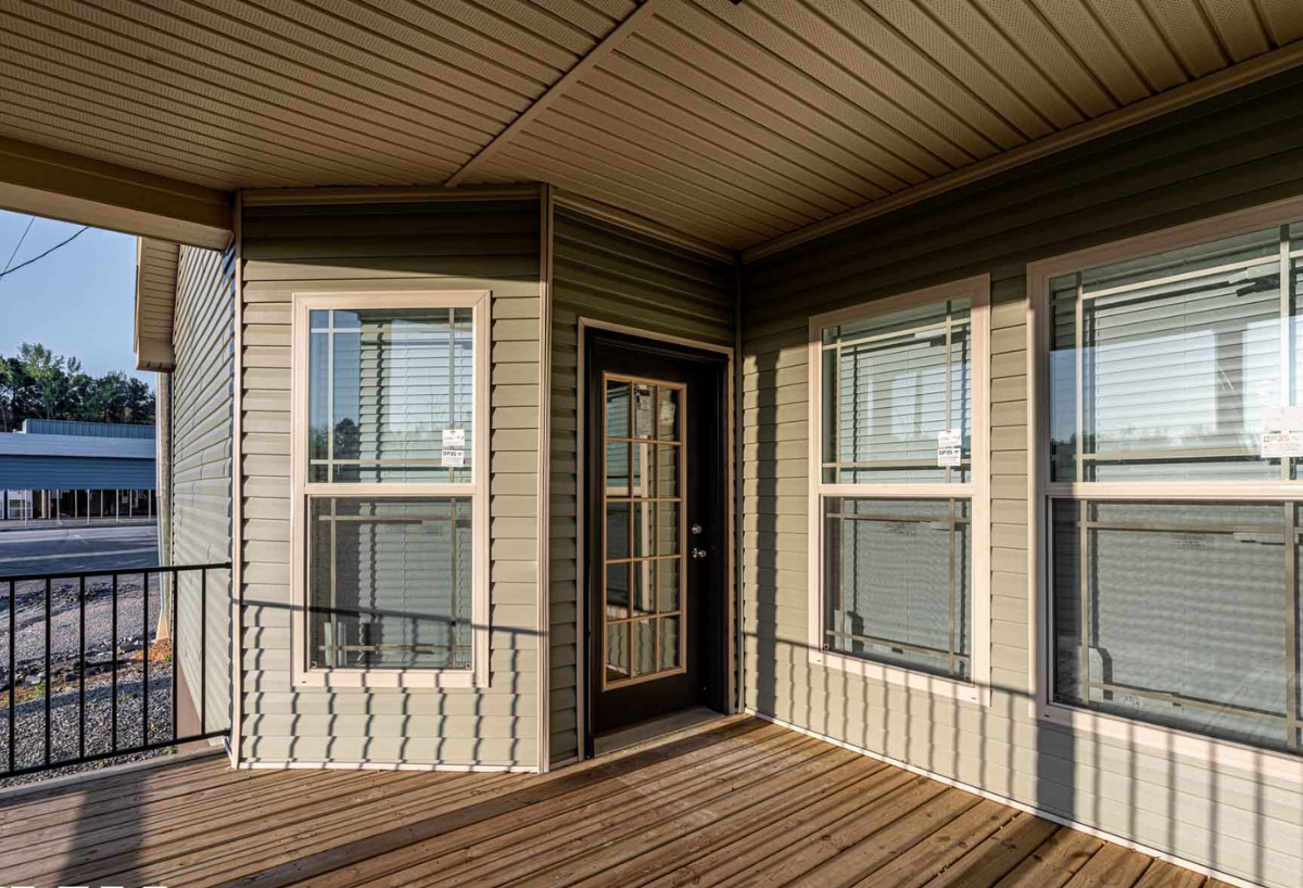 Covered porch with wooden floor and beige siding, featuring glass-paneled door and large windows. Shadows cast by railing create a serene atmosphere.