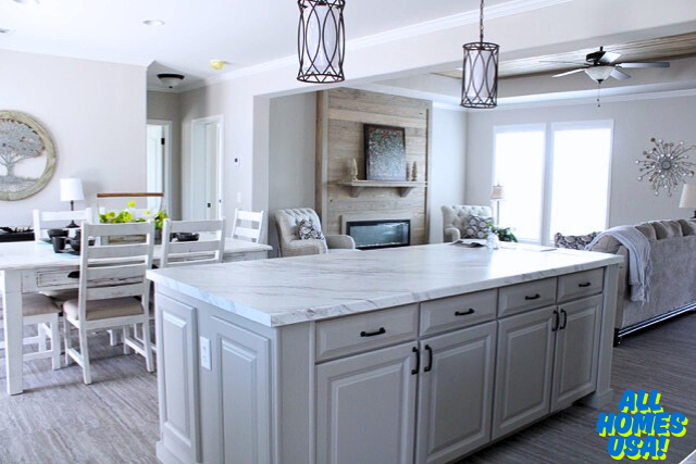 Bright kitchen and living area with a large marble island, white cabinets, and elegant pendant lights. Cozy seating by a fireplace and large windows. Image for illustrative purposes.