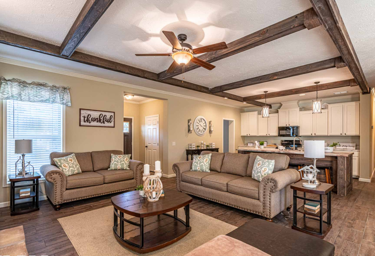 Cozy living room with brown sofas, patterned cushions, and dark wood coffee table. A ceiling fan and exposed beams add warmth. Kitchen visible in the background.