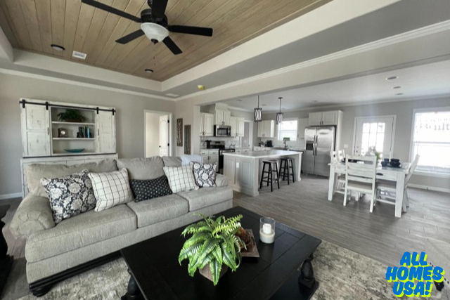 Cozy open-concept living room and kitchen with neutral tones. Sofa, patterned cushions, wooden coffee table with plant, island with stools, dining area. Image for illustrative purposes.