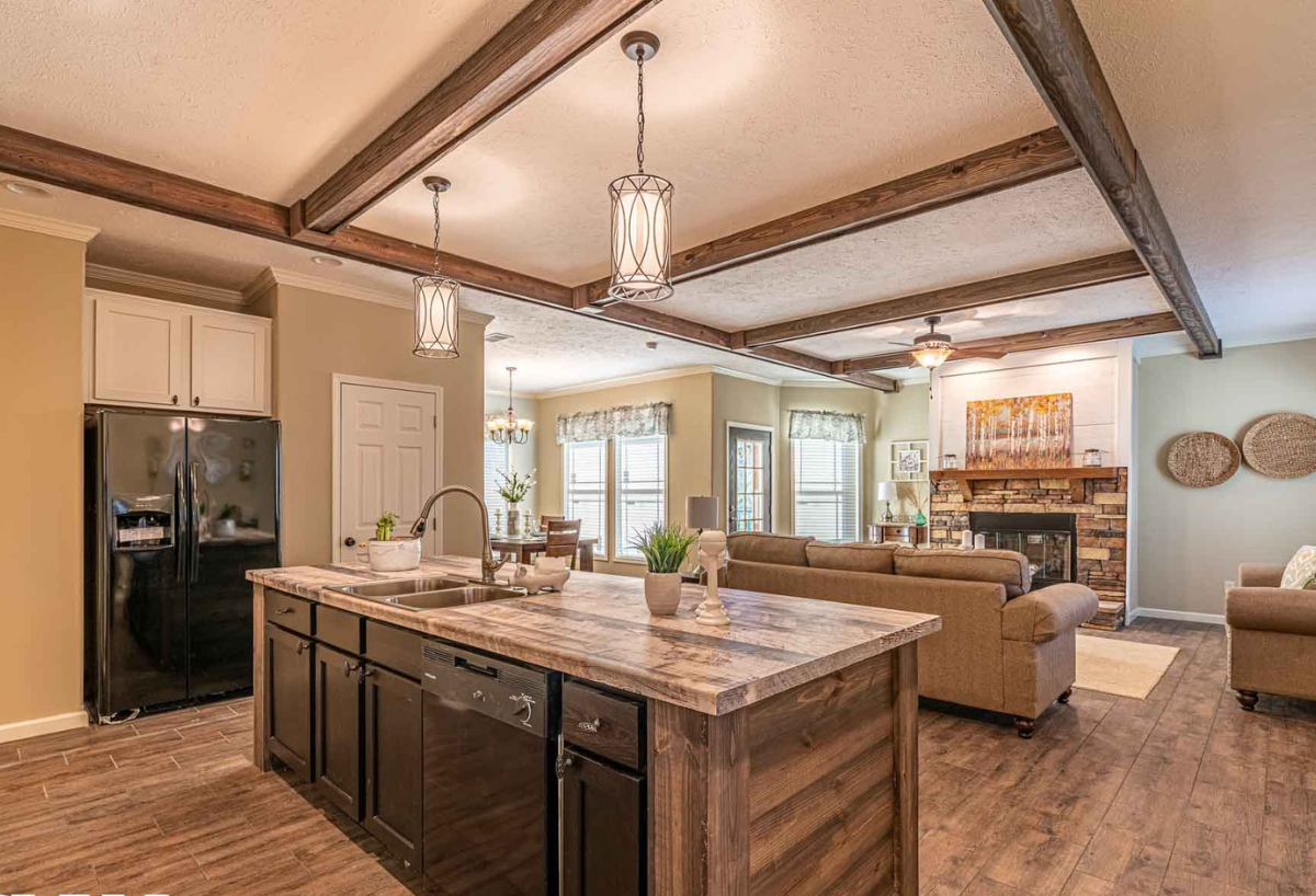 Warm, rustic kitchen and living area with wood beams. Central island with dark cabinets, modern lights, and plants. Cozy seating, fireplace in the background.