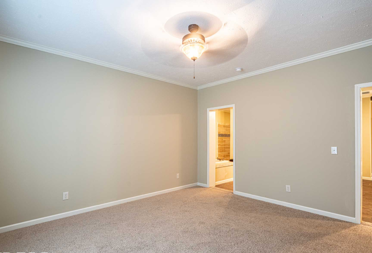An empty carpeted room features beige walls, a ceiling fan with light, and two doorways leading to a bathroom and hallway, creating a calm ambiance.