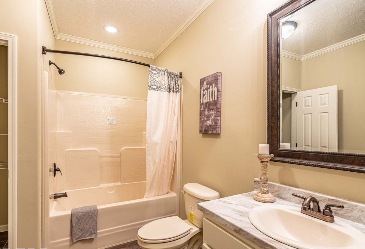 Warm-toned bathroom with a tub-shower combo, brown shower curtain, and a white vanity. A framed mirror reflects the door. A sign reads "faith."