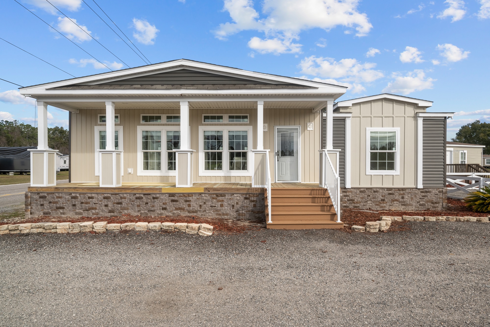 Single-story modular home with a beige facade, large windows, and a small porch. The roof is white, and the setting is under a bright blue sky.
