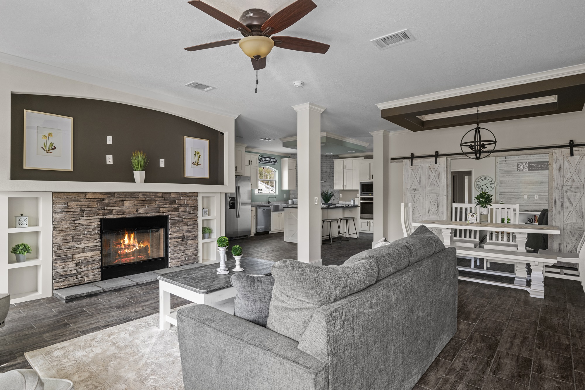 Stylish living room with gray sofa, stone fireplace, potted plants, and candles. Ceiling fan above, open kitchen, and farmhouse dining area visible. Cozy ambiance.