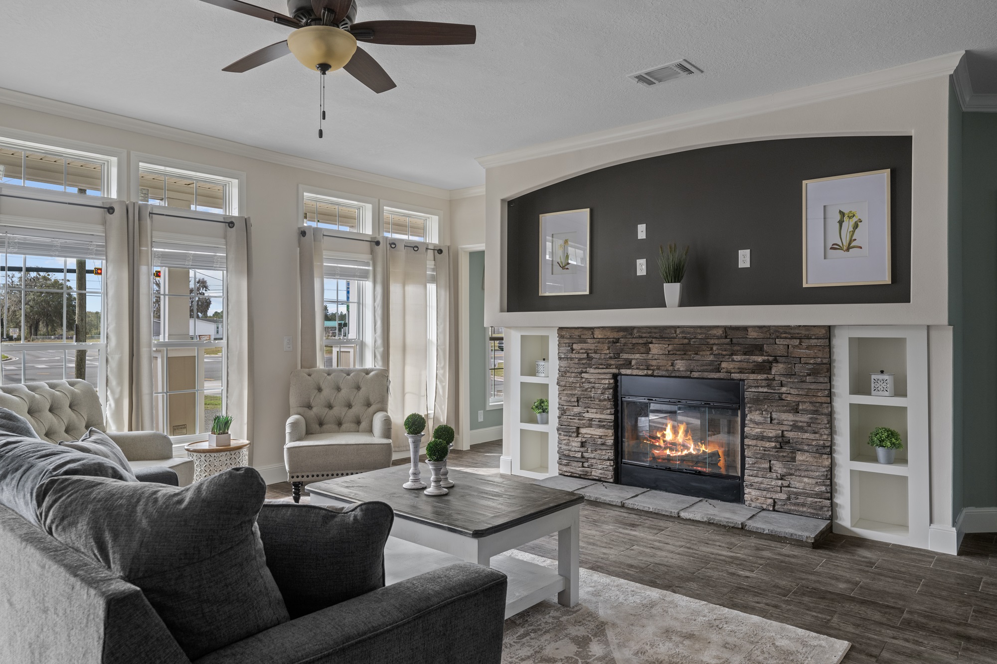 A cozy living room with large windows, a stone fireplace, and two armchairs. A gray sofa faces a wooden coffee table, with calm, neutral tones.