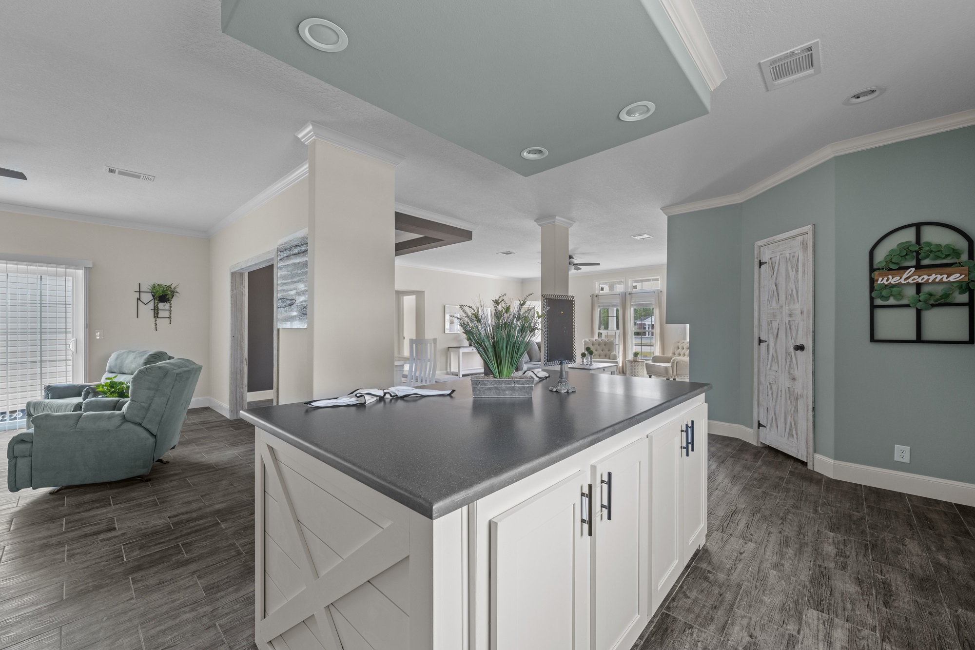 Modern open-plan living room and kitchen with gray flooring. Features a large island, light blue recliner, and "welcome" sign on a teal wall, conveying a cozy atmosphere.