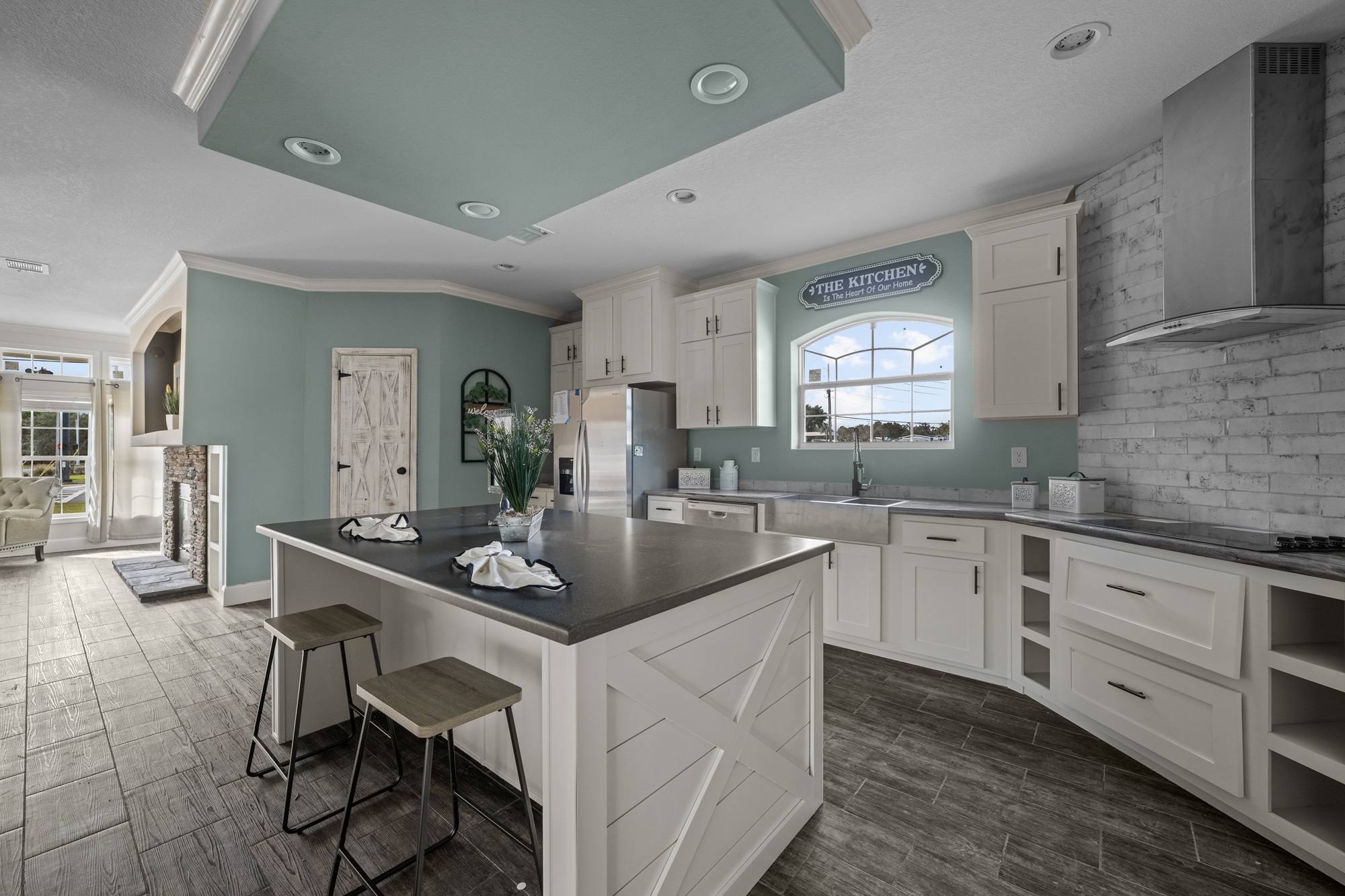 Modern kitchen with light teal walls, white cabinets, and gray countertops. A central island with stools faces a rustic door. Bright natural light.