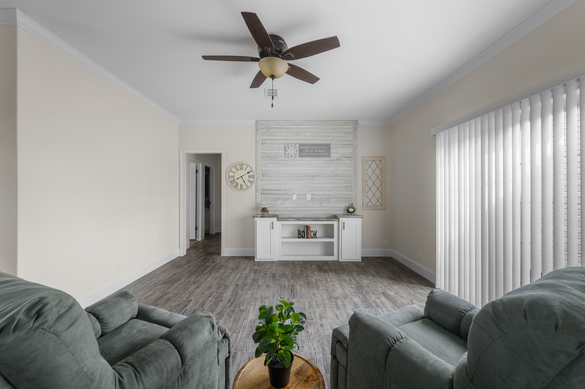 Bright living room with two sage green armchairs facing a small round table with a plant. A clock and framed art adorn a shiplap accent wall. Vertical blinds and ceiling fan included.