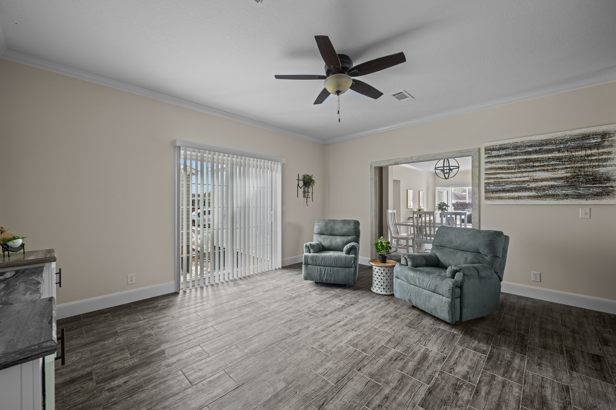 Minimalist living room with wood tile flooring, two teal recliners, ceiling fan, and large window with vertical blinds. Patio view and dining room visible.