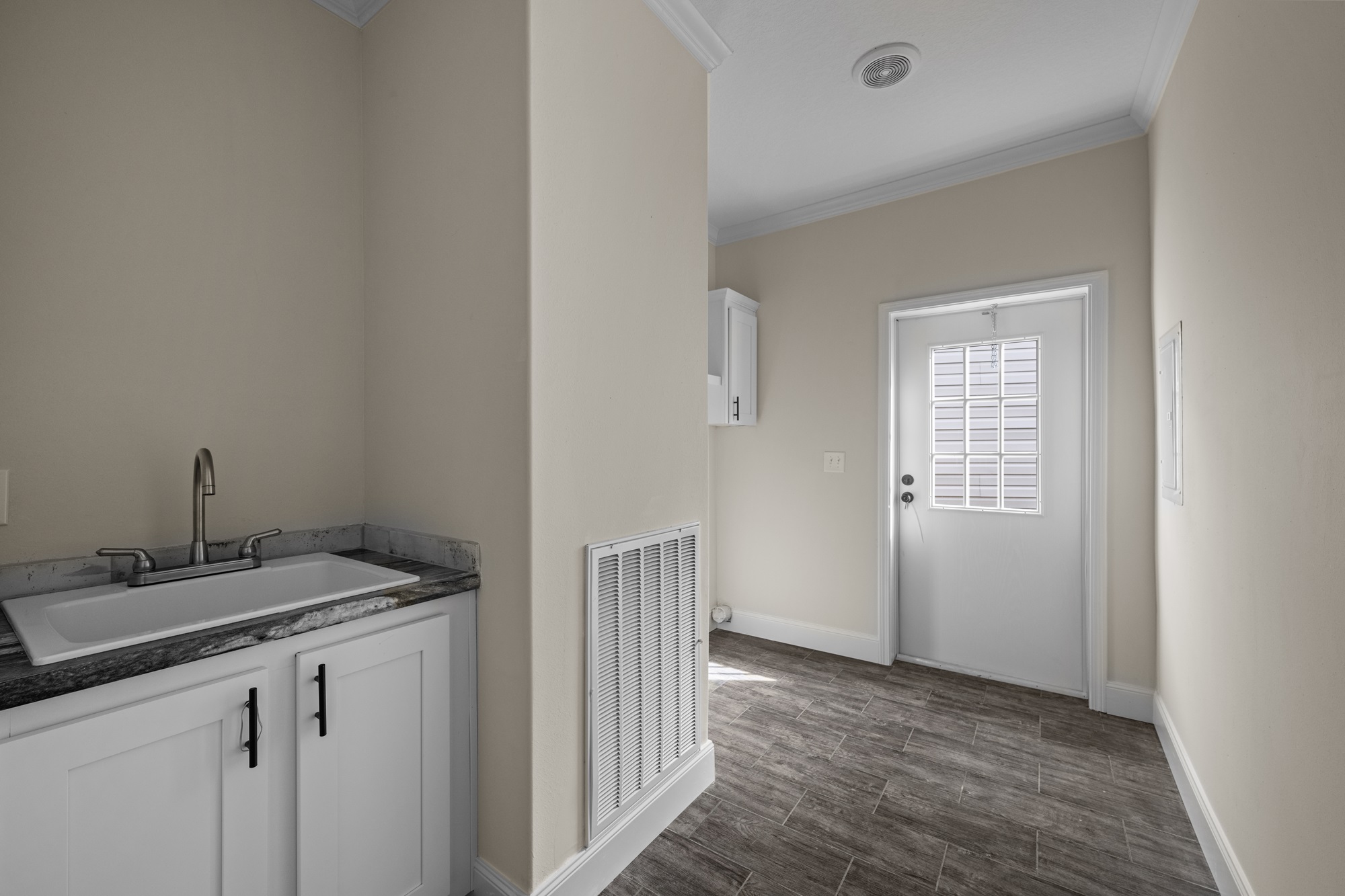 A bright, minimalistic laundry room with beige walls and dark wood flooring. Features a white sink with a granite countertop and a door with a window.