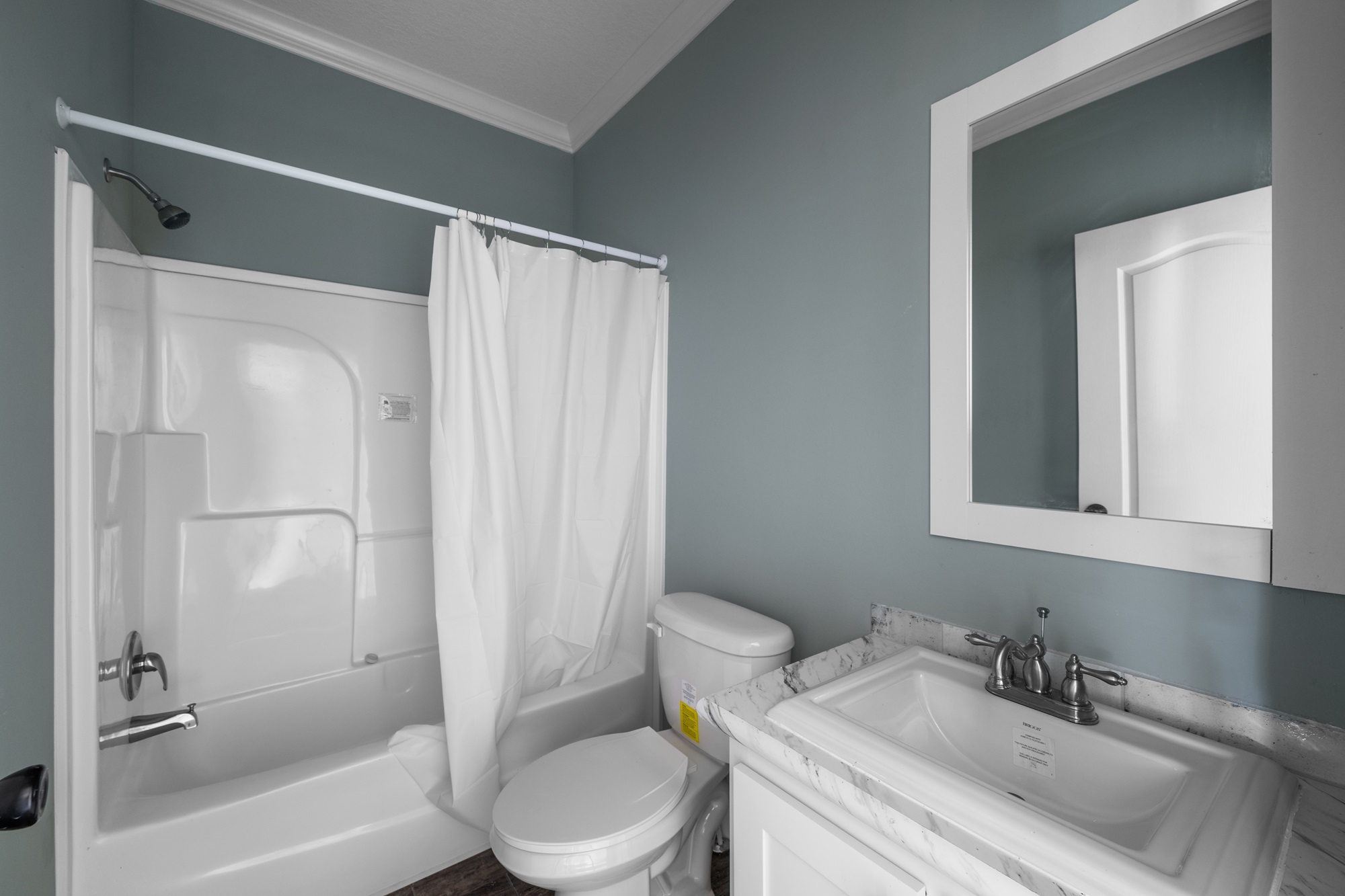 A small bathroom with blue-gray walls features a white bathtub and shower with a curtain, a toilet, and a sink with a mirror above, creating a clean, simple atmosphere.