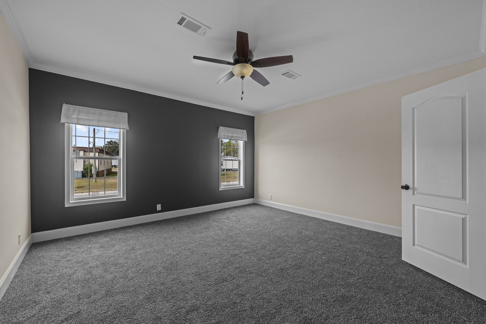 Spacious empty room with gray carpet, beige and dark gray walls, two windows, white door, and a ceiling fan. The atmosphere is calm and minimalist.
