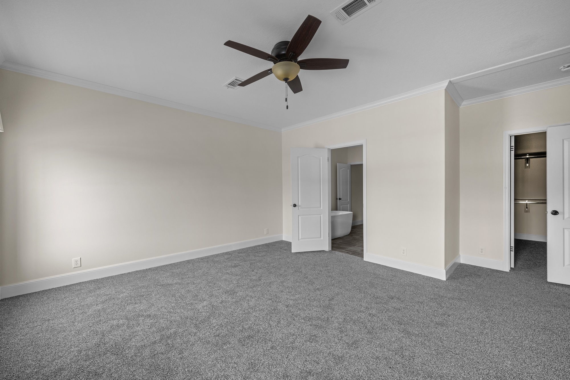 Spacious, empty room with gray carpet, beige walls, and a ceiling fan. Open doors reveal a bathroom with a freestanding tub and a walk-in closet.