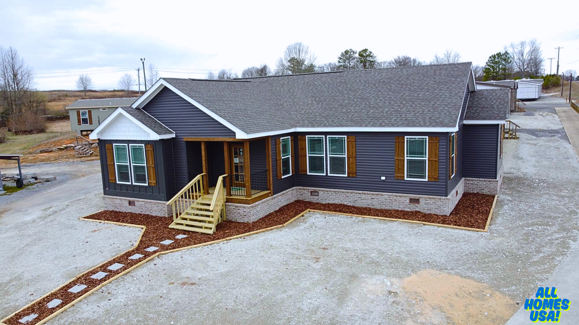 Modern, single-story dark gray house with wooden shutters and a small porch. Situated on a gravel lot with a neat path, conveying a calm rural setting. Image for illustrative purposes.
