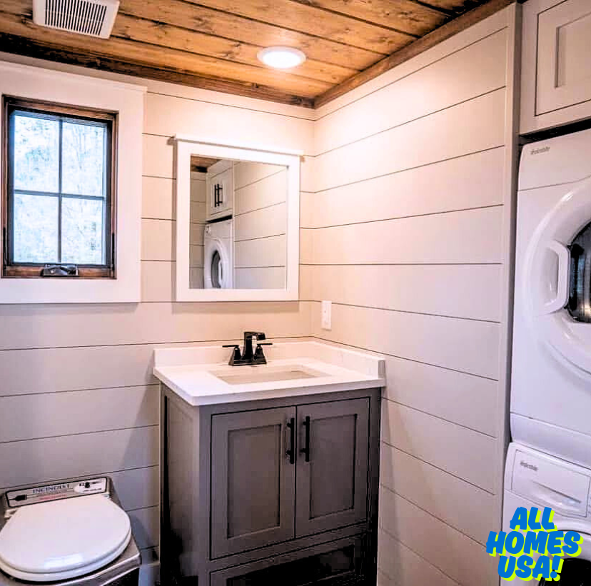 Cozy bathroom with wood ceiling, white shiplap walls, and gray vanity. Includes sink, mirror, toilet, small window, and a stackable washer-dryer unit. Image for illustrative purposes.