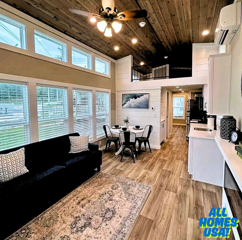Spacious tiny home interior with high ceilings, wood flooring, and natural light. Cozy dining area, modern kitchen, black sofa, calm ambiance. Image for illustrative purposes.