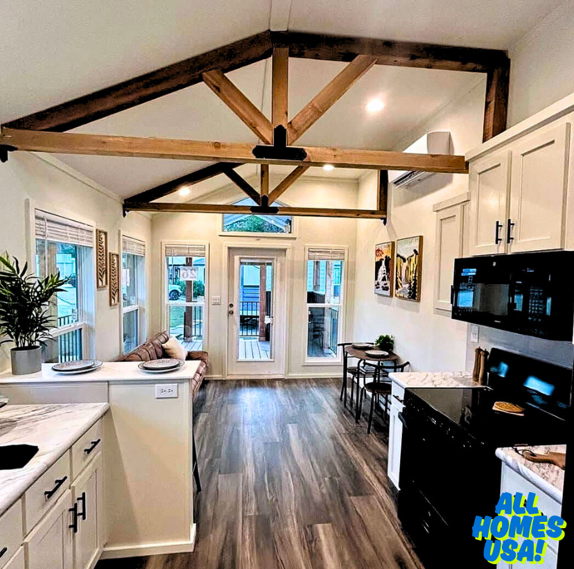 Modern kitchen and dining area with vaulted ceilings and wooden beams. Features sleek cabinets, a black stove, and large windows for natural light. Cozy seating area in a corner adds a welcoming touch. Image for illustrative purposes.