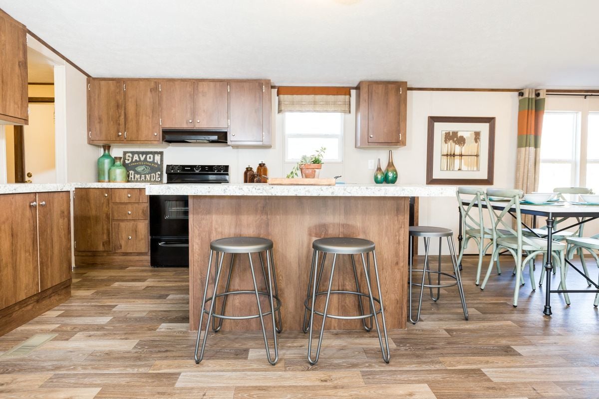 Bright, modern kitchen with wooden cabinets and flooring, featuring a central island with three metal stools. A cozy dining area is visible with a round table and green chairs, creating a warm and inviting atmosphere.